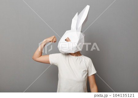 Image of strong unrecognizable woman wearing white t shirt and paper rabbit mask standing isolated over gray background, raised her arm, showing biceps and her power. Image of strong unrecognizable woman wearing white t shirt and paper rabbit mask standing isolated over gray background, raised her arm, showing biceps and her power. 104135121