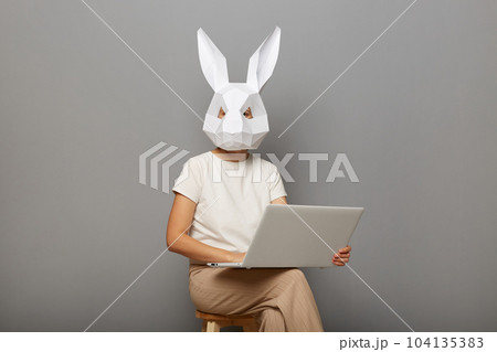 Portrait of anonymous female wearing rabbit mask and white t-shirt, sitting on chair with notebook in hands, freelancer working online, isolated over gray background. Portrait of anonymous female wearing rabbit mask and white t-shirt, sitting on chair with notebook in hands, freelancer working online, isolated over gray background. 104135383