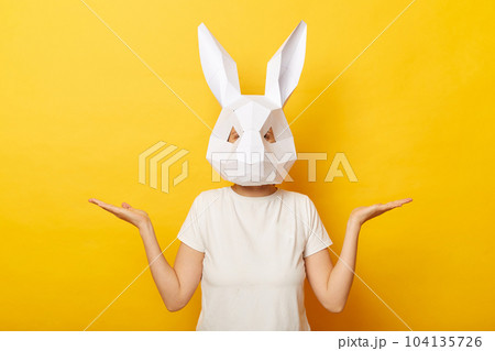 Horizontal shot of woman wearing white T-shirt and paper rabbit mask standing isolated over yellow background, posing with spread hands, presenting advertisement area on her palms. 104135726