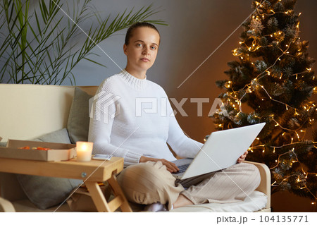 Indoor shot of calm attractive woman with bun hairstyle working on laptop computer sitting on sofa decorated for Christmas celebration, looking at camera, wearing white sweater. 104135771