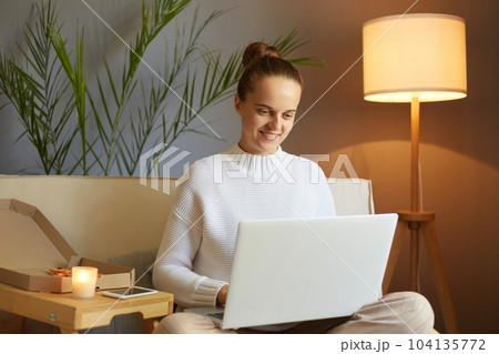 Indoor shot of smiling beautiful woman wearing white sweater sitting in cozy living room and working on laptop, having online job, female freelancer sitting on sofa in front of notebook. 104135772