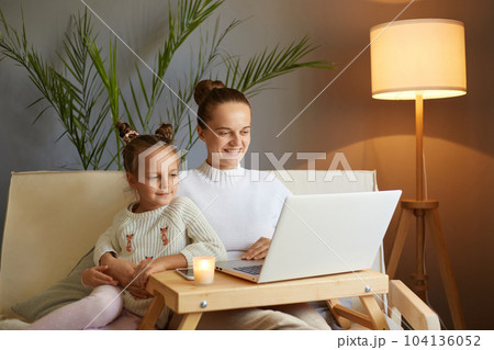 Horizontal shot of smiling woman with her cute little daughter, wearing white sweater sitting on sofa and watching videos or cartoons, spending time together. Horizontal shot of smiling woman with her cute little daughter, wearing white sweater sitting on sofa and watching videos or cartoons, spending time together. 104136052