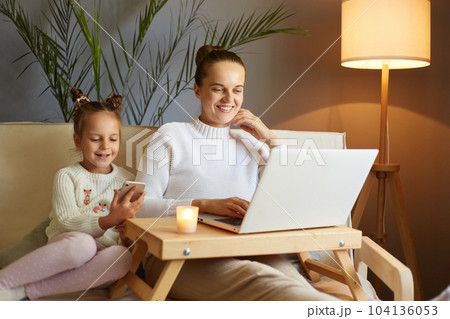 Indoor shot of smiling happy mother and daughter siting on the couch and using laptop, little girl holding mobile phone, family spending time together, expressing happiness. Indoor shot of smiling happy mother and daughter siting on the couch and using laptop, little girl holding mobile phone, family spending time together, expressing happiness. 104136053