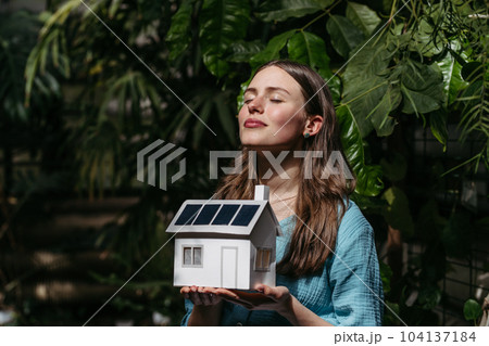 Young woman in jungle holding paper model of house with solar panels, concept of renewable energy and protection of nature. Young woman in jungle holding paper model of house with solar panels, concept of renewable energy and protection of nature. 104137184