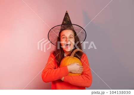 Indoor shot of excited amazed Caucasian woman dressed in witch hat holding orange pumpkin isolated on neon light background, extremely happy female celebrating halloween. 104138030