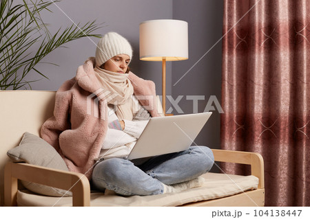 Side view portrait of calm sad depressed woman wearing cap and winter coat sitting on the sofa at home, holding laptop, looking at display with disappointed expressing, fells cold and bored. 104138447