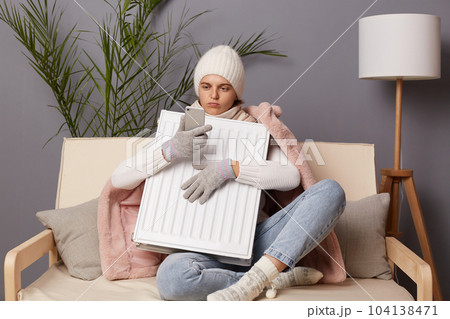 Portrait of Caucasian woman in coat and hat sit in cold living room embracing electric heating to get warm and using cell phone, call to service, broken central heating. 104138471