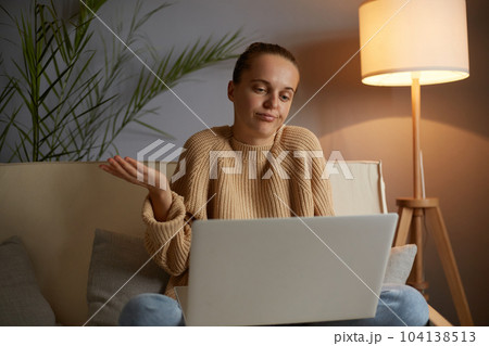 Indoor shot of puzzled confused Caucasian woman wearing beige jumper and jeans sitting on cough in lotus pose, having video call on laptop, shrugging shoulders, looks helpless. Indoor shot of puzzled confused Caucasian woman wearing beige jumper and jeans sitting on cough in lotus pose, having video call on laptop, shrugging shoulders, looks helpless. 104138513