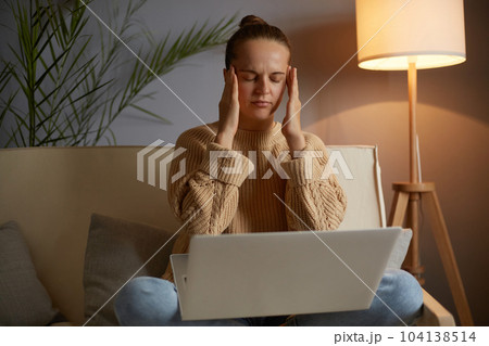 Portrait of sad depressed tired sick woman freelancer wearing beige jumper and jeans sitting on cough, massaging her temples, suffering headache, keeps eyes closed. 104138514