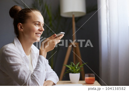 Indoor shot of smiling joyful woman with bun hairstyle sitting at table near window, using mobile phone for recording voice messages, smiling happily. 104138582