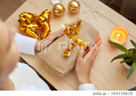 Top view of faceless female holding in hands christmas gift with gold bow on background of table with christmas ribbons and balls, preparing for New Year holidays. Merry Christmas. Top view of faceless female holding in hands christmas gift with gold bow on background of table with christmas ribbons and balls, preparing for New Year holidays. Merry Christmas. 104138619