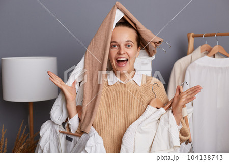 Photo of excited amazed extremely happy woman with clothes on her head holding clothing on hangers, standing with funny look, screaming happily, rejoicing to buy new attires. 104138743