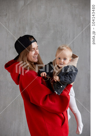 Young cute long haired mom in red hoodie and black baseball cap is holding and hugging her cute baby in beige hoodie and funny motorcycle jacket. Studio shooting, gray background. Young cute long haired mom in red hoodie and black baseball cap is holding and hugging her cute baby in beige hoodie and funny motorcycle jacket. Studio shooting, gray background. 104143480