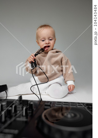 A cute baby in a beige hoodie sits on the floor and plays with DJ headphones and a DJ mixing console. Music and fun. Isolated on a gray background. A cute baby in a beige hoodie sits on the floor and plays with DJ headphones and a DJ mixing console. Music and fun. Isolated on a gray background. 104143484