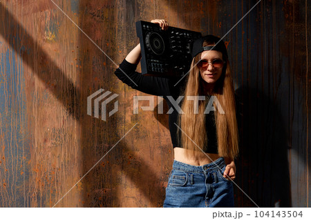 A young smiling long-haired DJ girl in a black top, jeans, a black baseball cap and sunglasses stands against a grunge brown wall and holds a black DJ mixing console in her hand. Music and beauty. 104143504