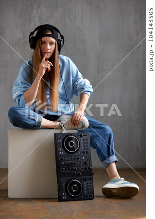A young pretty long-haired DJ girl in a blue sweater, jeans, a black baseball cap and black headphones sits with a DJ mixing console. Studio shot, gray background. 104143505