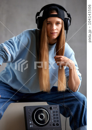 A young pretty long-haired DJ girl in a blue sweater, jeans, a black baseball cap and black headphones sits with a DJ mixing console. Studio shot, gray background. 104143506