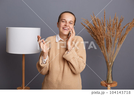 Portrait of smiling attractive positive woman wearing warm sweater standing against gray wall with lamp and dried flower on background, talking on cell phone, expressing happiness. Portrait of smiling attractive positive woman wearing warm sweater standing against gray wall with lamp and dried flower on background, talking on cell phone, expressing happiness. 104143718