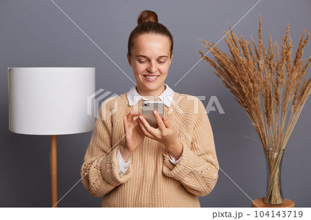 Image of joyful woman wearing warm sweater using cell phone for checking cocial networks, expressing positive emotions, standing against gray wall with lamp and dried flower on background. Image of joyful woman wearing warm sweater using cell phone for checking cocial networks, expressing positive emotions, standing against gray wall with lamp and dried flower on background. 104143719
