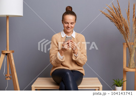 Portrait of good looking woman wearing beige jumper sitting on chair and using smart phone for checking social networks or e-mail, browsing internet, answering messages. Portrait of good looking woman wearing beige jumper sitting on chair and using smart phone for checking social networks or e-mail, browsing internet, answering messages. 104143741