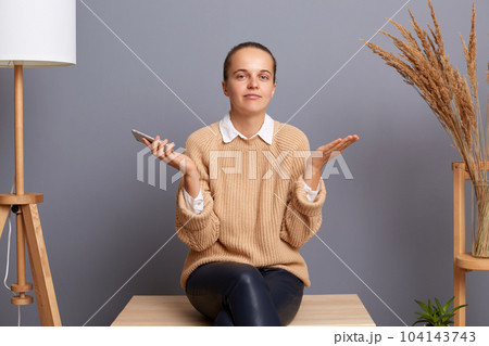 I don't know. Attractive confused woman wearing beige sweater sitting on chair with raised arms and looking at camera, holding mobile in hands, doesn't know answer. 104143743