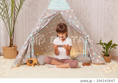 Indoor shot of shocked little girl with pigtails wearing white t shirt posing in wigwam at home and using phone, looking at display with open mouth, sees something astonishing in Internet. Indoor shot of shocked little girl with pigtails wearing white t shirt posing in wigwam at home and using phone, looking at display with open mouth, sees something astonishing in Internet. 104143847