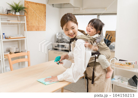ダイニングで背負い紐で子供をおんぶしてテーブルを拭いている若い女性 104150213