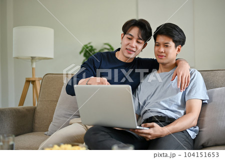 A happy Asian gay man enjoys watching a movie through a laptop with his boyfriend on a couch A happy Asian gay man enjoys watching a movie through a laptop with his boyfriend on a couch 104151653
