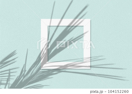 Natural light casts shadows from the palm branch on square frame of white paper lying on a blue facture background. Mock up with an overlay of plant shadows 104152260