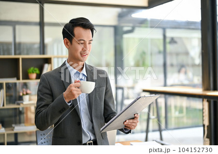 A handsome Asian businessman reading business report and sipping coffee in the office. 104152730