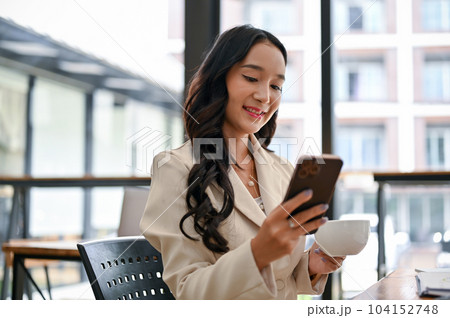 A happy Asian businesswoman using her smartphone and sipping coffee on her lunch break 104152748