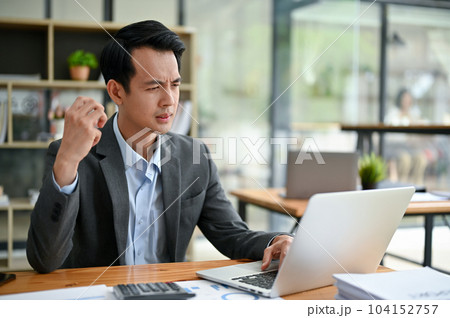 A thoughtful Asian businessman focuses on his tasks on his laptop, working at his desk A thoughtful Asian businessman focuses on his tasks on his laptop, working at his desk 104152757