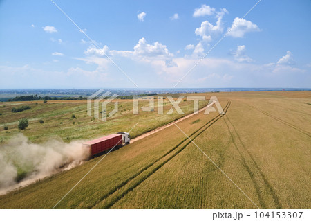 Aerial view of cargo truck driving on dirt road between agricultural wheat fields making lot of dust. Transportation of grain after being harvested by combine harvester during harvesting season Aerial view of cargo truck driving on dirt road between agricultural wheat fields making lot of dust. Transportation of grain after being harvested by combine harvester during harvesting season 104153307