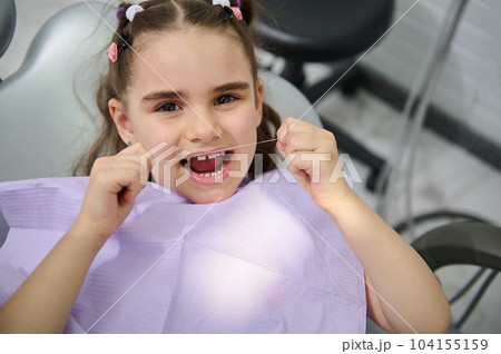 Close-up portrait of beautiful little girl using dental floss, brushing her teeth, sitting in the dentist's chair and smiling cutely, looking at the camera. Oral hygiene concept for caries prevention Close-up portrait of beautiful little girl using dental floss, brushing her teeth, sitting in the dentist's chair and smiling cutely, looking at the camera. Oral hygiene concept for caries prevention 104155159