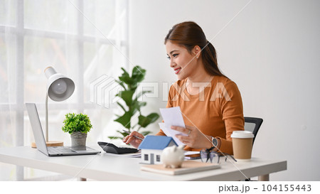 A beautiful Asian woman sits at her desk, paying her bills online on her laptop. 104155443