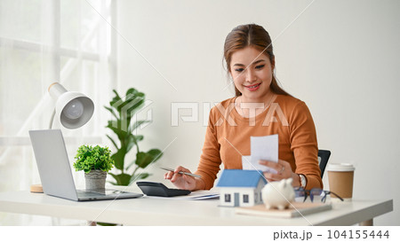 An attractive Asian woman sits at a table, checking her bills, calculating monthly expenses 104155444