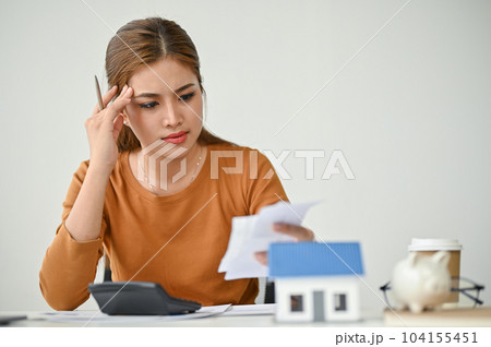 A stressed Asian woman sits at a table, checking her bills, calculating monthly expenses 104155451