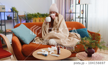 Sick ill woman suffering from cold or allergy sitting on home sofa sneezes wipes snot into napkin 104156697