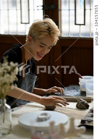 A happy Asian gay man artist kneading raw clay to sculpt an ornament in his studio. A happy Asian gay man artist kneading raw clay to sculpt an ornament in his studio. 104157055