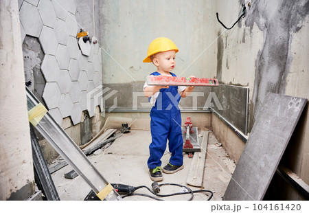 Boy construction worker holding spirit level instrument. Kid in safety helmet and work overalls standing in apartment bathroom. Home renovation and childhood concept. 104161420