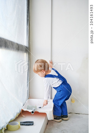 Little boy construction worker standing by the window and painting white wall in apartment. Kid in work overalls using paint brush while playing at home under renovation. Little boy construction worker standing by the window and painting white wall in apartment. Kid in work overalls using paint brush while playing at home under renovation. 104161493