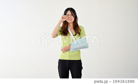 A sad woman with her accidentally broken arm in an arm sling is crying and wiping her tears 104161890