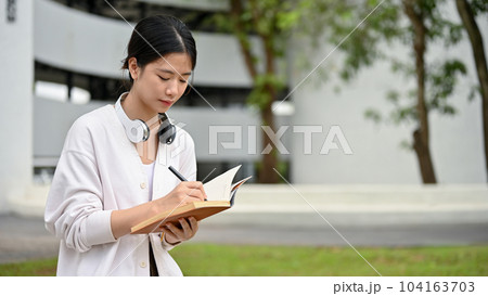 A focused Asian female student is in the campus park, writes her diary, sketches the view A focused Asian female student is in the campus park, writes her diary, sketches the view 104163703