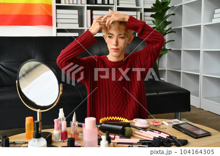 Young gay man doing daily makeup in living room, applying base foundation on face with sponge. LGBTQ, lifestyle and makeup concept Young gay man doing daily makeup in living room, applying base foundation on face with sponge. LGBTQ, lifestyle and makeup concept 104164305
