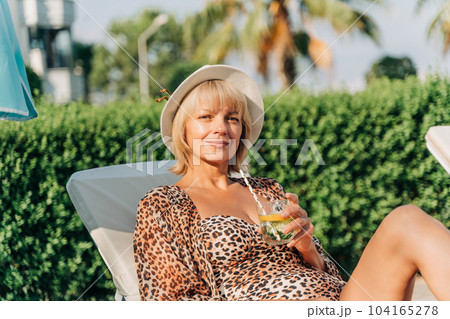 Portrait of happy mature adult woman relaxing deck chair near the pool and looking at camera. Beautiful caucasian senior woman in hat drink lemonade cocktail enjoying summer vacation 104165278