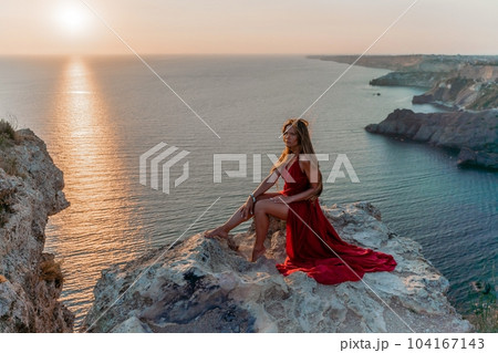 Woman sunset sea red dress, side view a happy beautiful sensual woman in a red long dress posing on a rock high above the sea on sunset. Woman sunset sea red dress, side view a happy beautiful sensual woman in a red long dress posing on a rock high above the sea on sunset. 104167143