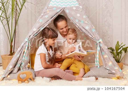 Indoor shot of mother with two little girls sitting together in wigwam, mom with her children reading interesting book with fairy tale in peetee tent. 104167504