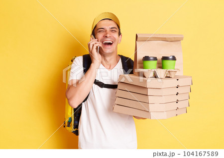 Indoor shot of happy laughing man courier with thermo backpack in white T-shirt and cap isolated over yellow background, talking phone, holding boxes with pizza and takeaway coffee. 104167589