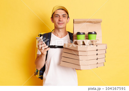 Horizontal shot of serious calm man courier with thermo backpack in white T-shirt and cap, looking at camera, holding client's order, standing with smart phone in hands isolated over yellow background Horizontal shot of serious calm man courier with thermo backpack in white T-shirt and cap, looking at camera, holding client's order, standing with smart phone in hands isolated over yellow background 104167591