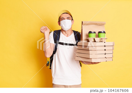 Photo of delivery man in yellow cap, white t-shirt and protective face mask holding pizza and coffee, clenched fists, rejoicing, posing isolated on yellow background 104167606